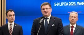 Wiceminister Rzymkowski stoi i mówi do mikrofonu na stojaku, za nim dwóch mężczyzn w garniturach, w tle niebieska ściana.