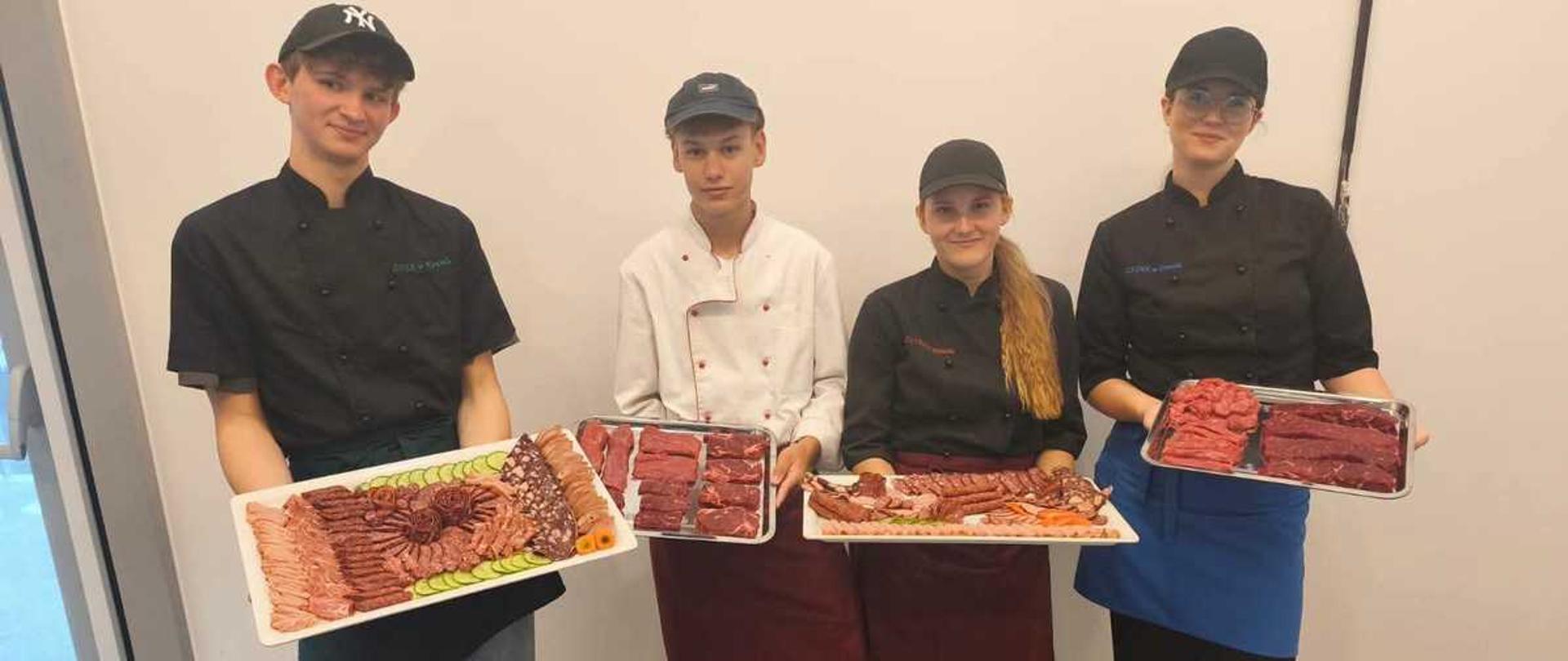 "Zdjęcie przedstawia czworo młodych ludzi – dwóch chłopaków i dwie dziewczyny – stojących obok siebie w jednym rzędzie na tle gładkiej, jasnej ściany. Wszyscy są ubrani w profesjonalne stroje kucharskie lub rzeźnickie (bluzy kucharskie, fartuchy oraz czapki z daszkiem) i prezentują trzymane w dłoniach tace z różnymi rodzajami mięsa oraz wędlin.
Oto szczegółowy opis osób od lewej do prawej:
• Pierwszy chłopak od lewej – uśmiecha się, ma na sobie czarną bluzę kucharską, ciemnozielony fartuch wiązany w pasie, luźne jeansy oraz czarną czapkę z daszkiem z białym logo New York Yankees. W rękach trzyma dużą, prostokątną białą tacę, na której estetycznie ułożono rzędy pokrojonych wędlin, kiełbas oraz boczku, ozdobione na brzegu plasterkami ogórka.
• Drugi chłopak – patrzy prosto w obiektyw, ubrany jest w białą bluzę kucharską z czerwonymi lamówkami, bordowy fartuch oraz ciemną czapkę z daszkiem. Trzyma podłużną metalową tacę wypełnioną surowymi, grubymi stekami wołowymi o intensywnie czerwonej barwie.
• Pierwsza dziewczyna od prawej strony chłopaka – ma długi blond warkocz opadający na ramię, nosi czarną bluzę, bordowy fartuch, czarne spodnie i czarną czapkę. W dłoniach trzyma szeroką tacę z różnorodnymi wyrobami mięsnymi, m.in. długimi kiełbaskami i kabanosami.
• Druga dziewczyna (ostatnia po prawej) – nosi okulary, czarną bluzę kucharską, jaskrawoniebieski fartuch, czarne spodnie oraz czarną czapkę. Prezentuje metalową tacę, na której znajdują się kawałki surowego, czerwonego mięsa (prawdopodobnie wołowiny).
Wszyscy stoją na ciemnosarej wykładzinie. Po lewej stronie kadru widoczny jest fragment przeszklonych drzwi wejściowych, przez które wpada naturalne światło. Atmosfera na zdjęciu jest profesjonalna, a młodzież wygląda na dumną z prezentowanych produktów."