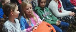 Na zdjęciu widoczne siedzące na krzesełkach dzieci dwoje z nich trzyma bojkę ratowniczą, koloru pomarańczowego. Dzieci poznają sprzęt ratowniczy podczas prowadzonych przez strażaków zajęć z zakresu bezpieczeństwa nad wodą w ramach programu "Aktywni Błękitni - szkoła przyjazna wodzie"