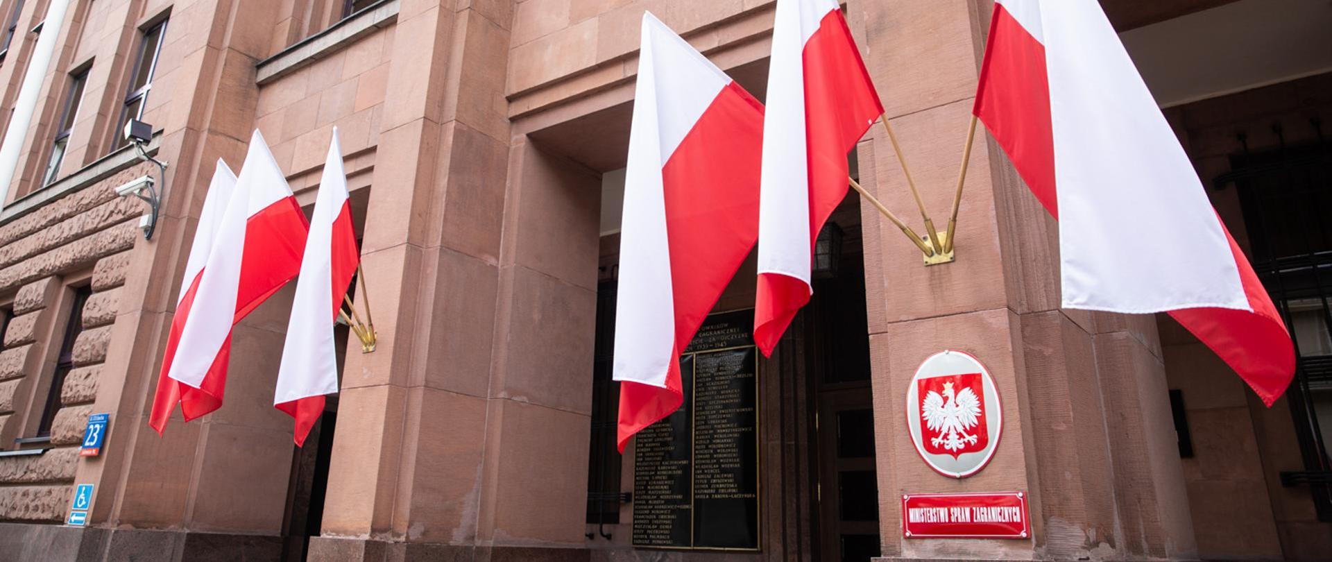 Na zdjęciu widać wejście do gmachu Ministerstwa Spraw Zagranicznych, wywieszone są biało-czerwone flagi, na kolumnie godło Polski oraz tabliczka z napisem Ministerstwo Spraw Zagranicznych.