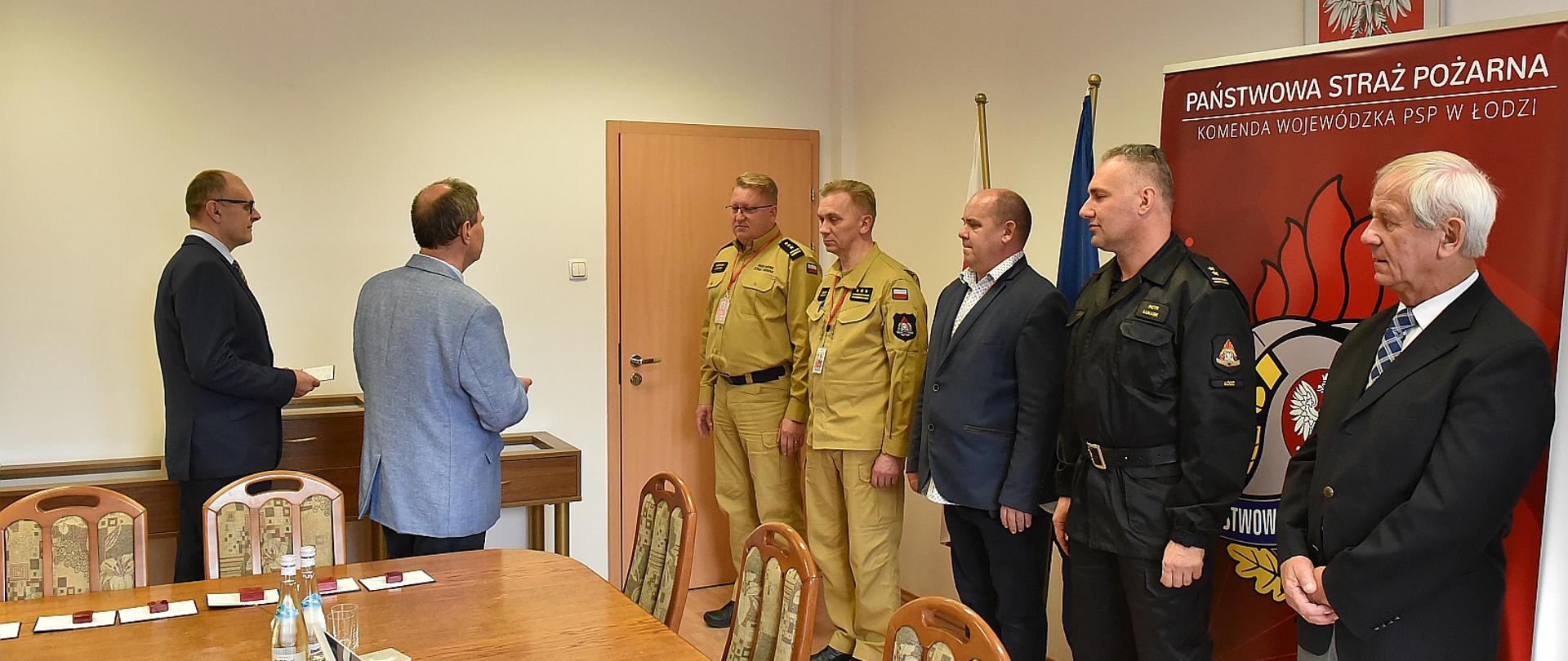 na zdjęciu siedem mężczyzn stojących w sali w tym trzech strażaków strażaków, dwóch w nowym umundurowaniu służbowym w kolorze piaskowych i jeden w umundurowaniu sztabowy w kolorze czarnym , pozostali ubrani są w marynarki