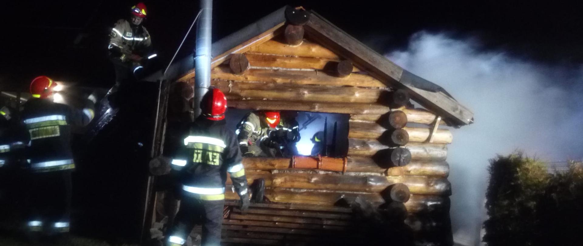 Zdjęcie wykonane w porze nocnej obrazuje budynek gospodarczy o konstrukcji drewnianej z widoczną spaloną częścią dachu. Widoczni strażacy usuwają spalone i nadpalone elementy konstrukcyjne obiektu i wyposażenia.
