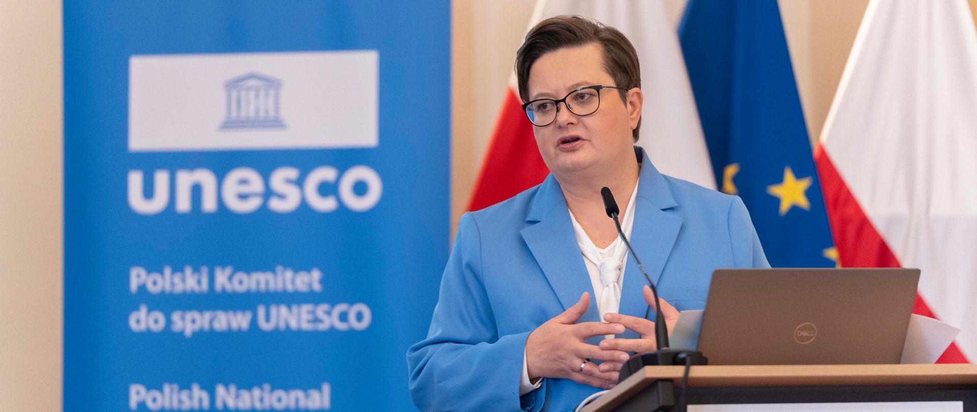 Kobieta (Katarzyna Lubnauer) w niebieskiej marynarce i okularach przemawia, gestykulując obiema rękami, stojąc za mównicą z laptopem. W tle widoczne są flagi Polski oraz Unii Europejskiej, a także niebieski rollup z logo UNESCO i napisem "Polski Komitet do spraw UNESCO".