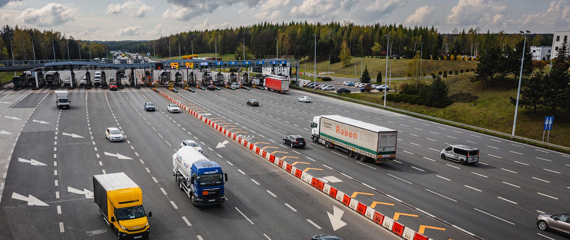 Widok z góry na wielopasmowy punkt poboru opłat na autostradzie A4. Przez bramki przejeżdżają samochody osobowe i ciężarowe, a na jezdni widoczne są liczne oznaczenia kierunkowe oraz separatory pasów ruchu. W tle znajduje się zalesiony teren i częściowo zachmurzone niebo. Na zdjęciu widoczna jest infrastruktura autostradowa związana z poborem opłat.