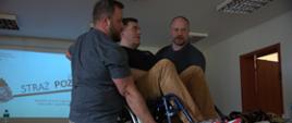 Dwie osoby podnoszą osobę siedzącą na wózku inwalidzkim podczas ćwiczeń w sali szkoleniowej; w tle znajduje się ekran z prezentacją oraz wyposażenie szkoleniowe.