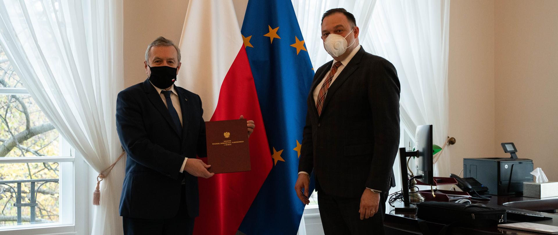 Minister Kultury, Dziedzictwa Narodowego i Sportu profesor Piotr Gliński wręcza nominację na nowego dyrektora COS Andrzejowi Kalinowskiemu. Premier i dyrektor stoją w gabinecie, w tle widoczne są flagi Polski i Unii Europejskiej.