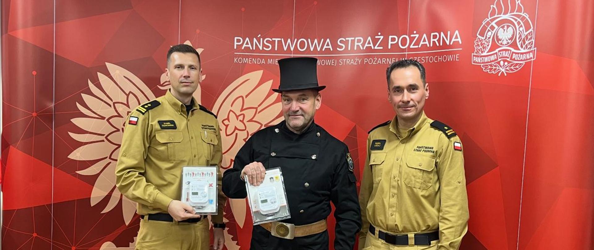 Zdjęcie przedstawia trzech mężczyzn stojących w rzędzie na tle czerwonej ściany z logotypem Państwowej Straży Pożarnej w Częstochowie. Jest to scena przedstawiająca przekazanie sprzętu. Dwaj mężczyźni po bokach ubrani są w żółto-brązowe mundury strażackie z dystynkcjami na pagonach. Mężczyzna po lewej trzyma w rękach małe, białe urządzenie w plastikowym opakowaniu, prawdopodobnie czujnik czadu lub dymu. Mężczyzna w centrum ubrany jest w tradycyjny czarny strój kominiarski, w tym czarną marynarkę, cylinder i ciemny pas ze złotą sprzączką, i on również trzyma w dłoniach podobne białe urządzenie w opakowaniu. Mężczyzna po prawej ubrany jest w analogiczny żółto-brązowy mundur służbowy. Wszyscy trzej mężczyźni patrzą prosto w obiektyw. Tło to czerwona ściana z dużym wzorem orła w koronie, a nad głowami mężczyzn widnieje napis: „PAŃSTWOWA STRAŻ POŻARNA KOMENDA MIEJSKA PAŃSTWOWEJ STRAŻY POŻARNEJ CZĘSTOCHOWIE”.