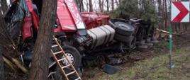  Na środku zdjęcia widać rozbity samochód ciężarowy z naczepą w kolorze czerwonym, który uderzył w drzewa i przewrócił się na prawy bok. Ciągnik siodłowy pojazdu ma uszkodzony cały przód, rozbitą przednią szybę, drzwi od strony kierowcy są otwarte, a przy nich oparta jest drabina nasadkowa w kolorze brązowym. Naczepa pojazdu leży w pozycji na prawym boku, nad którą zwisa nadłamana gałąź drzewa. W dolnej części zdjęcia widać pobocze, na którym rośnie trawa i znajdują się również uszkodzone elementy pojazdu. Po prawej stronie zdjęcia widnieje znak drogowy w kolorze biało czerwonym. Za uszkodzonym pojazdem widać rosnące iglaste drzewa.