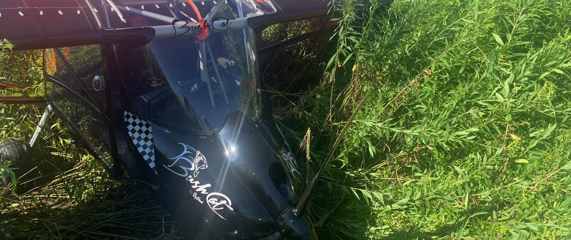 Na zdjęciu widać awaryjne lądowanie awionetki w trawie. Awionetka jest kolory czarnego, ogon koloru czerwonego ze złamanym przednim kołem, na pierwszym planie widoczne śmigło koloru czarnego. Kadłub samolotu leży na ziemi.