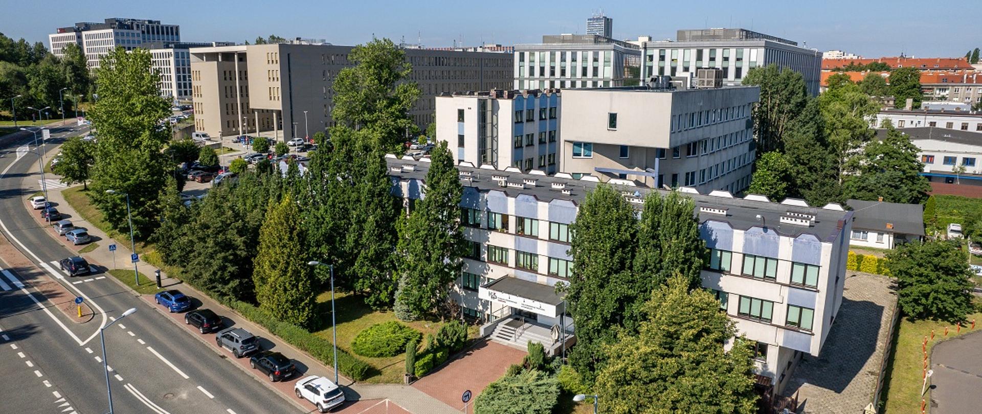 Słoneczny dzień, widok z ok. 30 m na kilkupiętowe biurowce, po lewej stronie widoczna droga, przy której stoją zaparkowane auta.