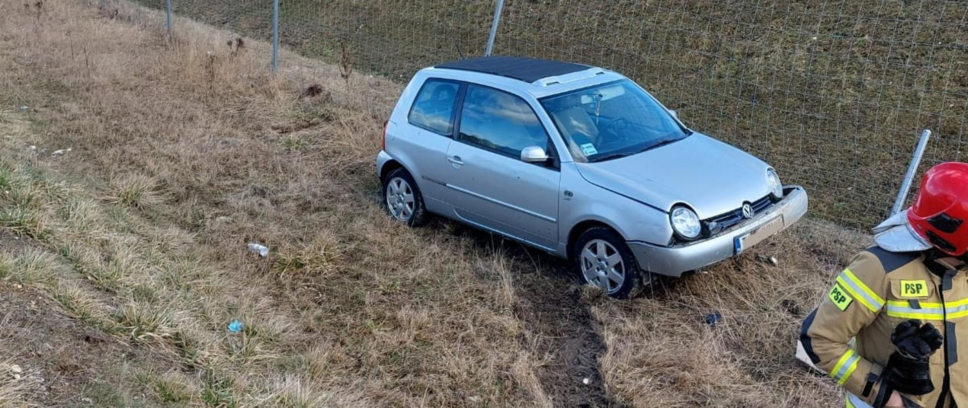 Na zdjęciu widać uszkodzony samochód osobowy znajdujący się poboczu drogi. Koło pojazdu widać strażaka w umundurowaniu specjalnym. 