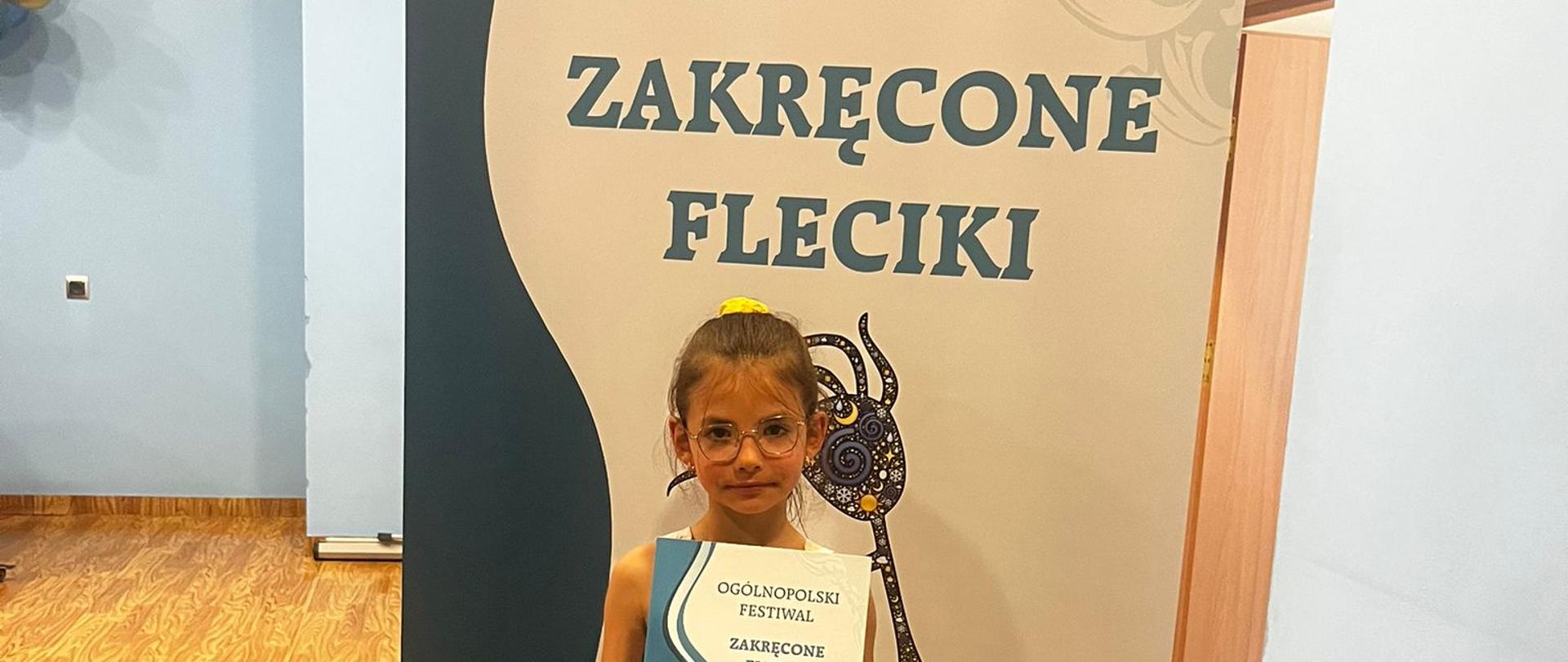 Zdjęcie przedstawia dziewczynkę z kolorowymi balonami i dyplomem laureata na tle plakatu Ogólnopolskiego Konkursu Zakręcone Fleciki