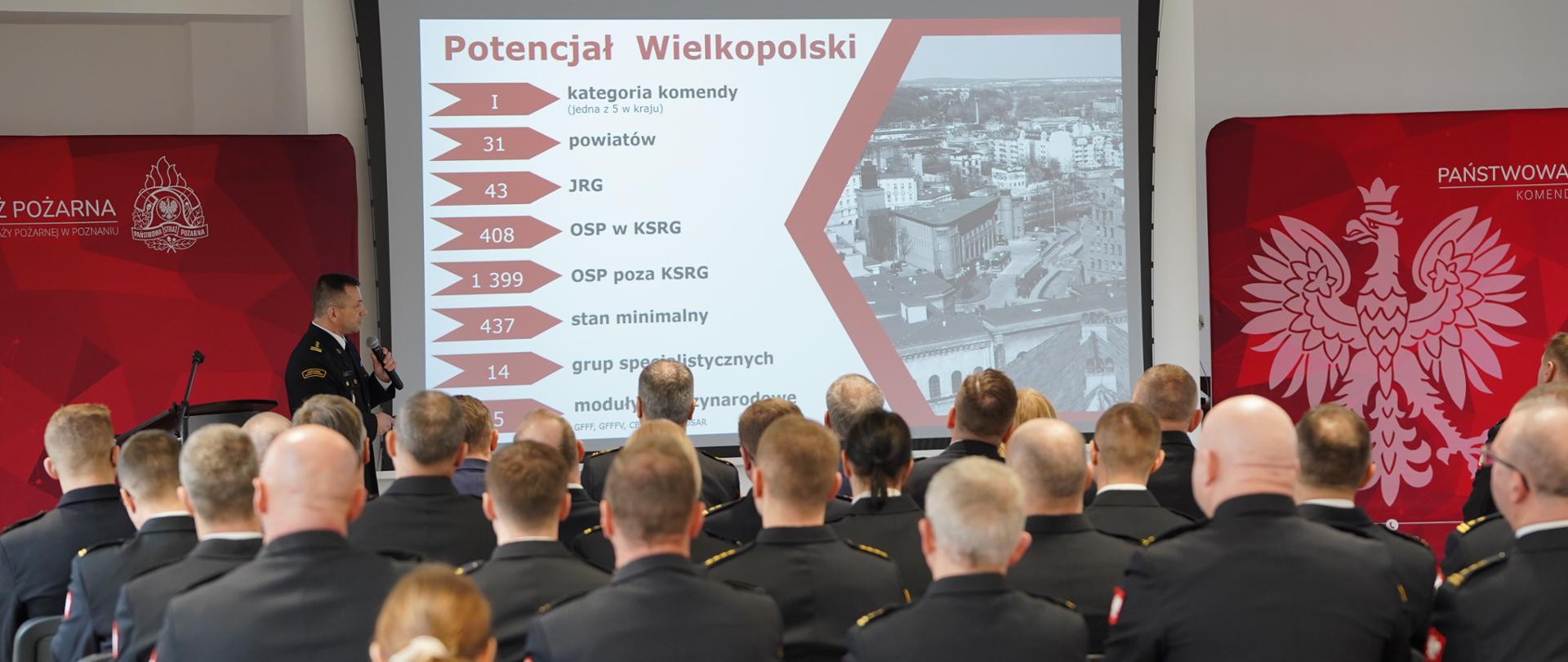  Sala konferencyjna z funkcjonariuszami Państwowej Straży Pożarnej uczestniczącymi w prezentacji. Na ekranie wyświetlana jest prezentacja na temat potencjału straży pożarnej w Wielkopolsce, a mówca w mundurze PSP przedstawia dane.