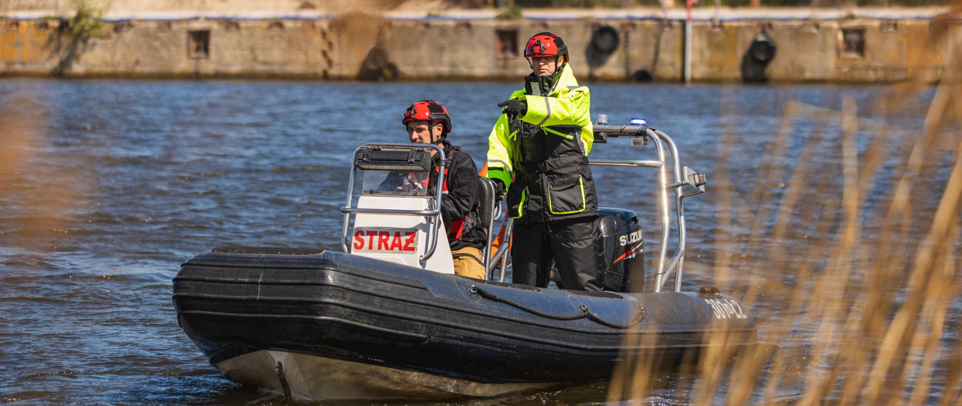 Strażacy w łodzi ratowniczej na wodzie; jeden z nich wskazuje kierunek ręką, drugi prowadzi jednostkę.