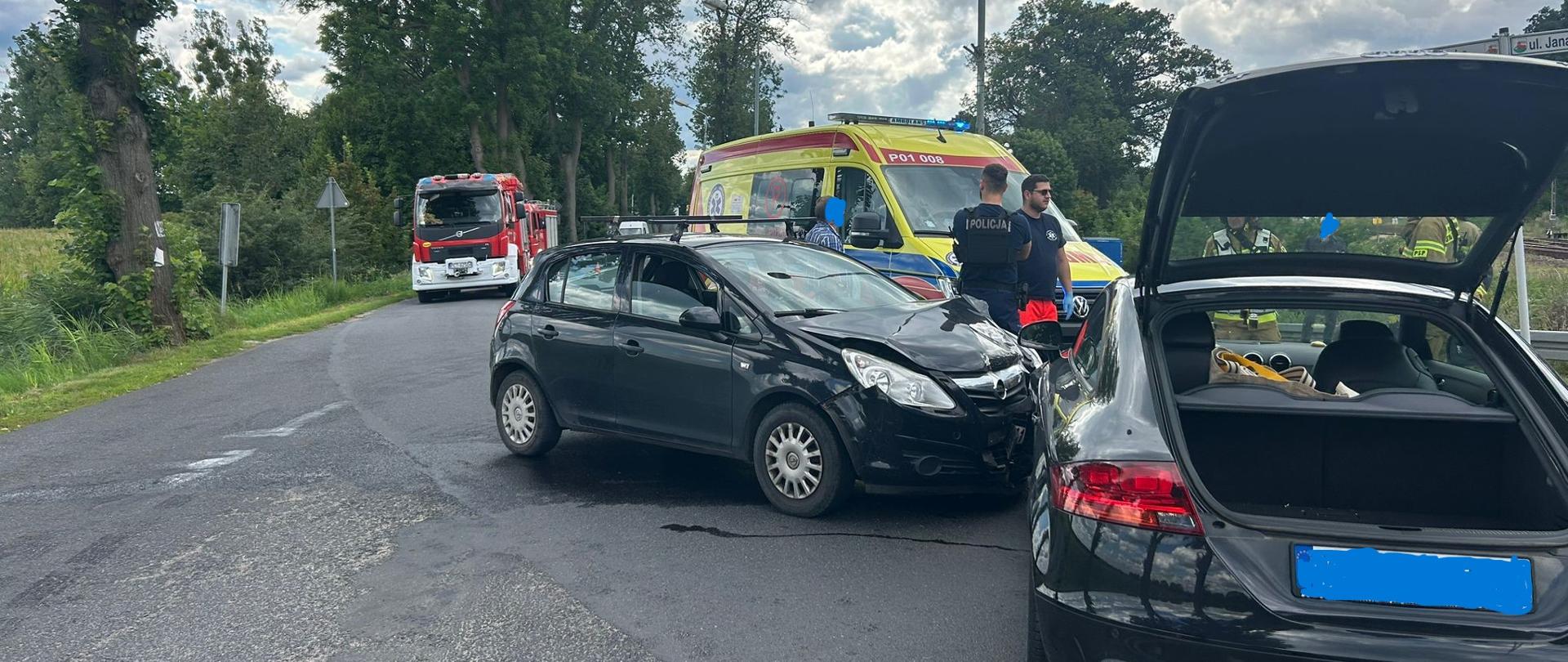 Na skrzyżowaniu doszło do kolizji dwóch samochodów osobowych, z widocznymi uszkodzeniami przodu jednego z nich. Obok stoją policjanci i ratownicy medyczni przy karetce pogotowia. W tle widoczny jest wóz strażacki.