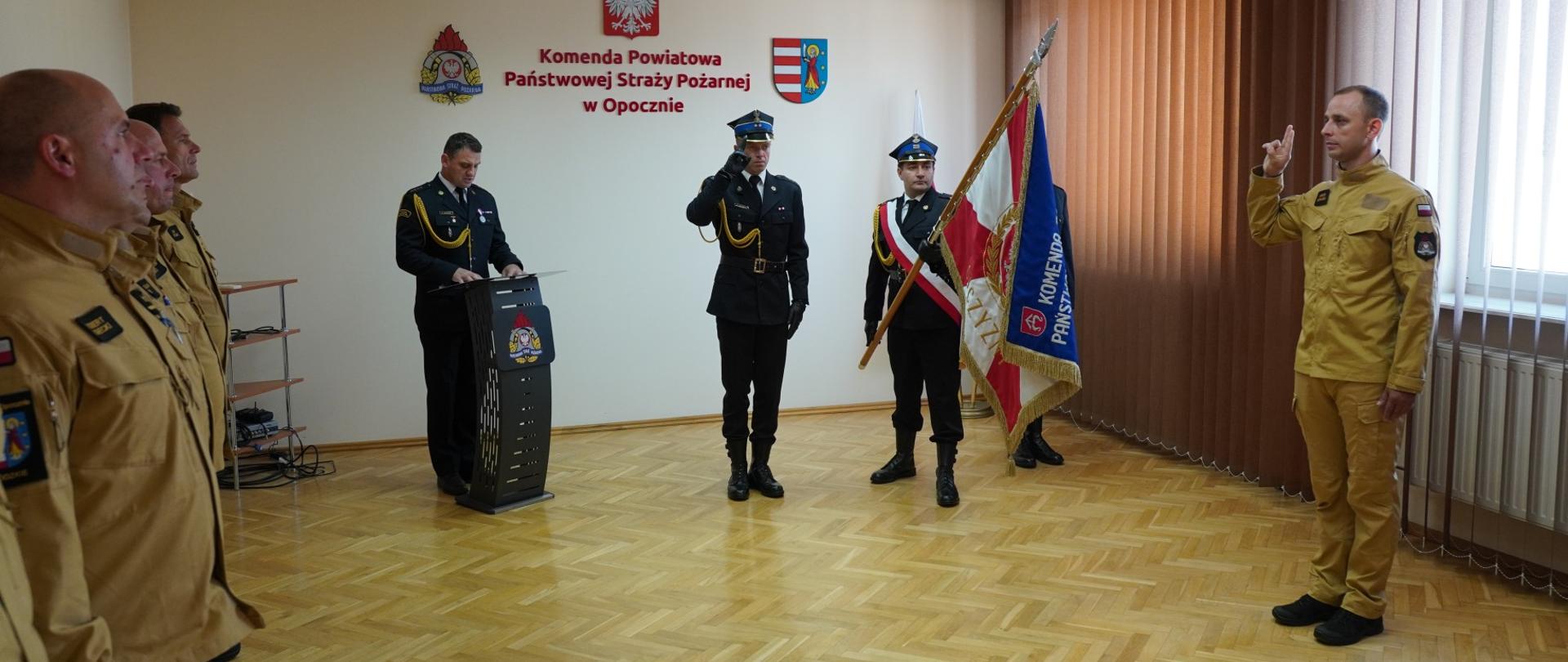 Sala konferencyjna. Strażak w ubraniu służbowym koloru pisakowego trzyma prawą dłoń zgiętą w łokciu i ślubuje w obecności pocztu sztandarowego oraz funkcjonariuszy PSP w Opocznie, w tle mównica Komendant Powiatowy w ubraniu galowym oraz napis Komendy Powiatowej PSP w Opocznie