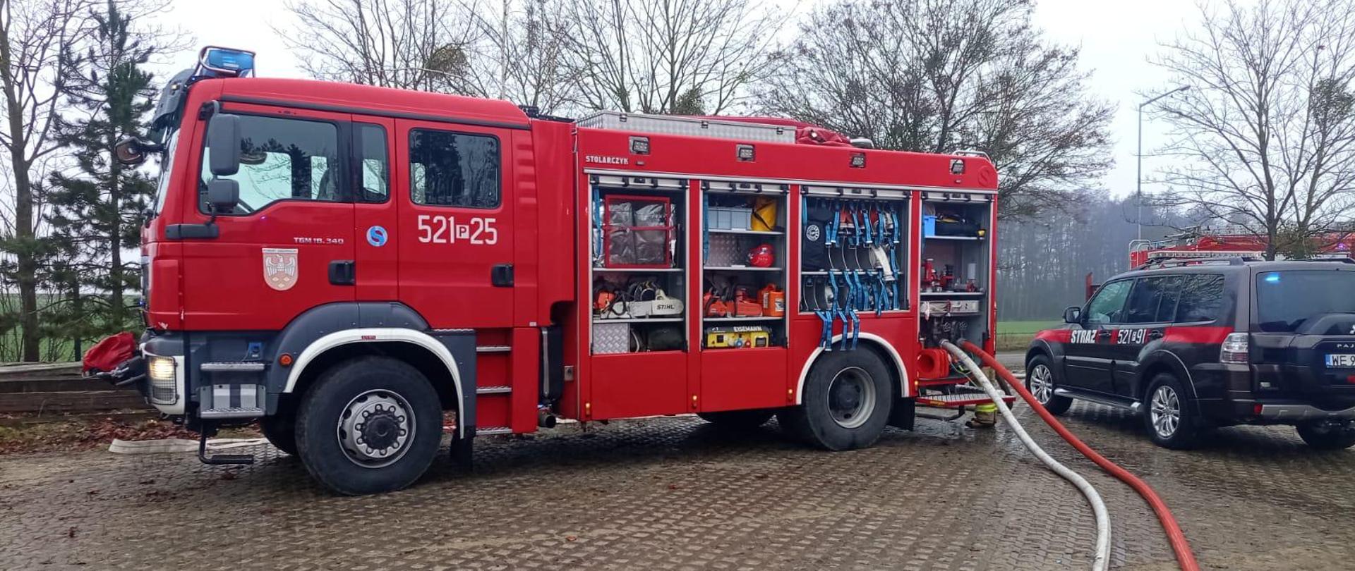 Wóz strażacki i osobowy samochód operacyjny stoją na utwardzonym placu podczas działań ratowniczych. Na ziemi rozłożone są węże gaśnicze. W tle widać drzewa. 