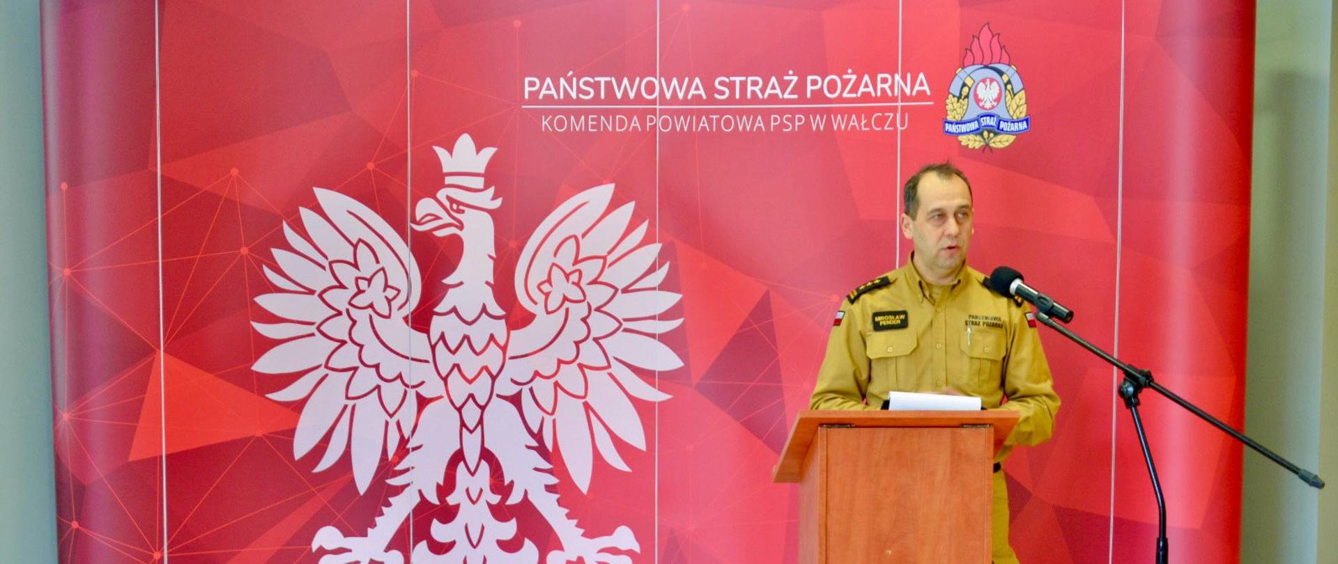Zachodniopomorski Komendant Wojewódzki państwowej Straży Pożarnej st. bryg. Mirosław Pender podczas okolicznościowego przemówienia. W tle widoczna jest czerwona ścianka medialna z kolorowym logo PSP. Zdjęcie powiększy się po kliknięciu na nie.