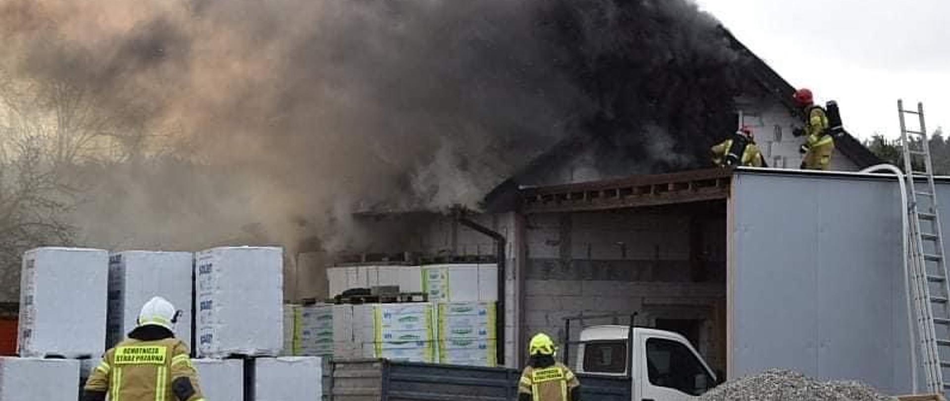 Pożar budynku gospodarczego. Pali się poddasze, na przybudówce 2 strażaków, przed budynkiem stoi samochód dostawczy i ułożone pustaki oraz styropian. Na pierwszym planie 2 strażaków 