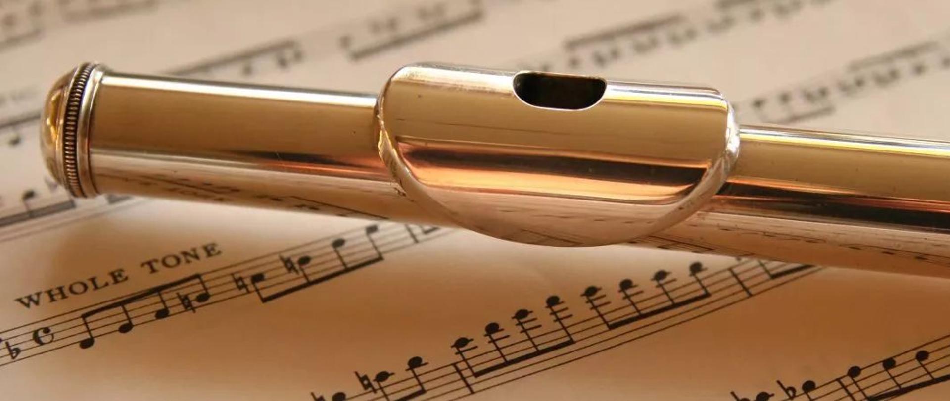 Zdjęcie. Fragment fletu (główka z ustnikiem) na tle nut.