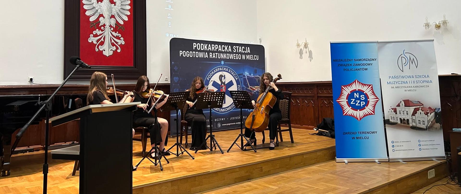 Cztery uczennice grają na skrzypcach, altówce i wiolonczeli na scenie Sali Królewskiej podczas Forum Eksperckiego, w tle banery Podkarpackiej Stacji Pogotowia Ratunkowego i PSM w Mielcu.