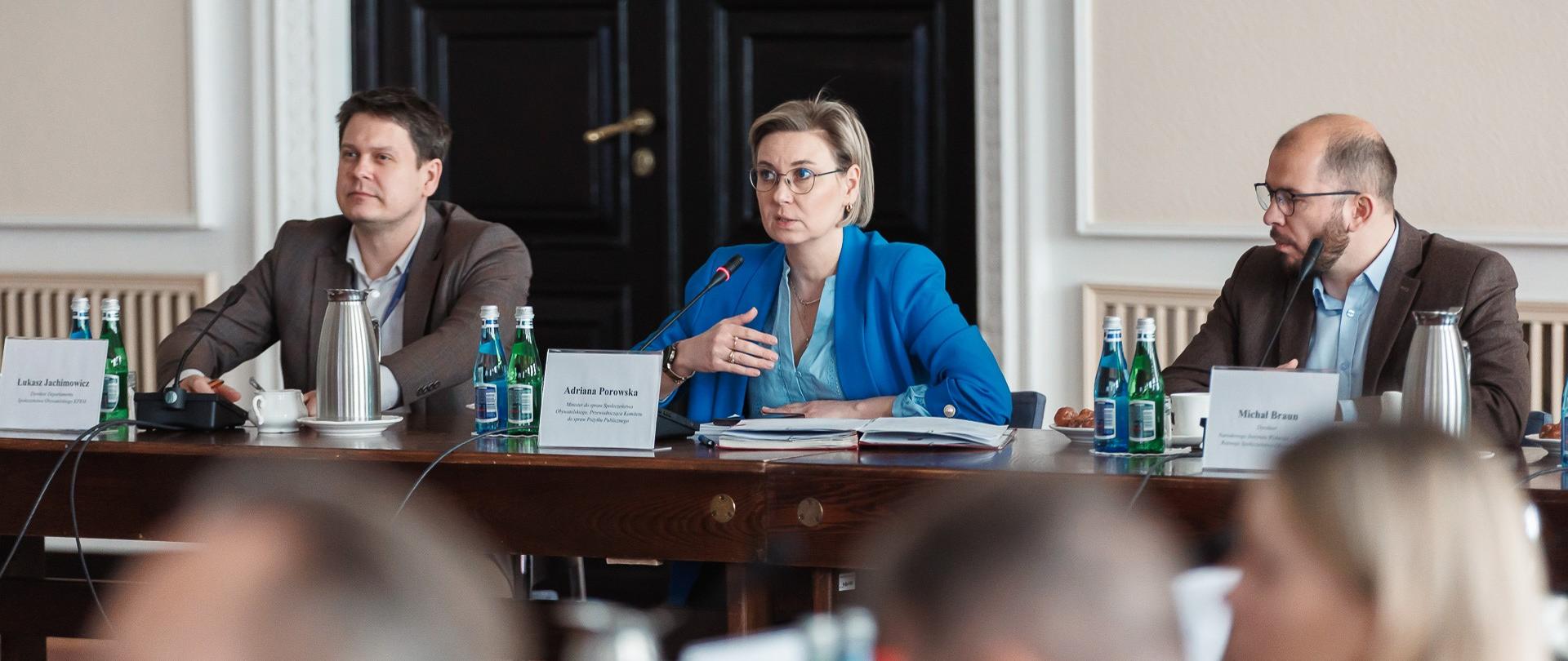 Zdjęcie przedstawia trzy osoby siedzące obok siebie przy długim stole konferencyjnym. Od lewej widoczny Łukasz Jachimowicz, Dyrektor Departamentu Społeczeństwa Obywatelskiego KPRM, Adriana Porowska, Ministra do spraw Społeczeństwa Obywatelskiego i Michał Braun, dyrektor Narodowego Instytutu Wolności. Ministra Porowska gestykuluje mówiąc do mikrofonu. Na pierwszym planie widoczne rozmyte sylwetki innych uczestników spotkania.