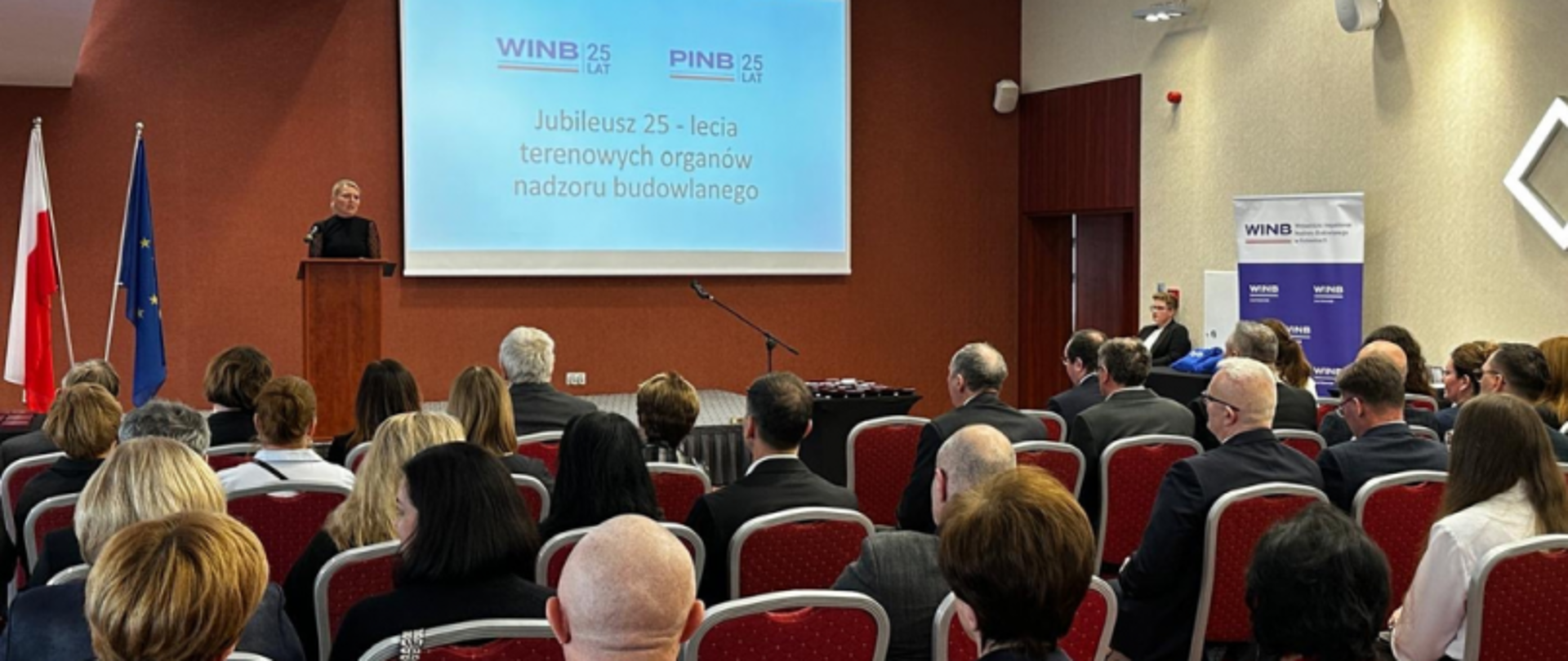 Sala konferencyjna z uczestnikami, prelegent przy mównicy, ekran z napisem o jubileuszu 25-lecia WINB i PINB.