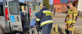 Zdjęcie przedstawia dwóch strażaków oraz kobietę na wózku inwalidzkim. Po lewej stronie widać auto do przewozu osób niepełnosprawnych.