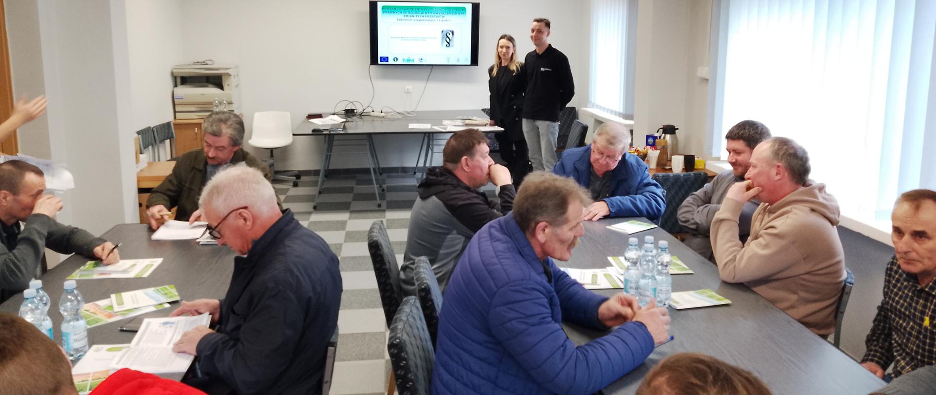 Grupa osób siedzi przy stołach, w głębi wyświetlony slajd, po jego prawej stronie stoją dwie osoby