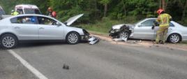 2 samochody osobowe po zderzeniu znajdujące się na jezdni, w tle służby ratownicze.
