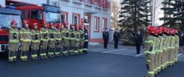 Przed kutnowską strażnicą odbywa się uroczysty apel związany z awansem na stanowisko Komendanta Powiatowego PSP w Łęczycy, wieloletniego z-cy Komendanta Powiatowego PSP w Kutnie bryg. Marcina Adamiaka. Przed strażnicą w umundurowaniu specjalnym stoją strazacy na baczność. W tle widać strażaków w mundurach wyjściowych. Za nimi na maszcie wciągnięta Flaga Polski. Jest pogodnie.