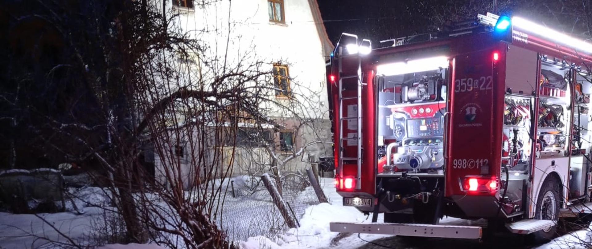 Zdjęcie przedstawia samochód pożarniczy stojący na drodze przy budynku objętym pożarem. W samochodzie otwarte są skrytki, w których znajdują się urządzenia i sprzęt ratowniczy. Skrytki są oświetlone. Zdjęcie wykonane po zmierzchu. 