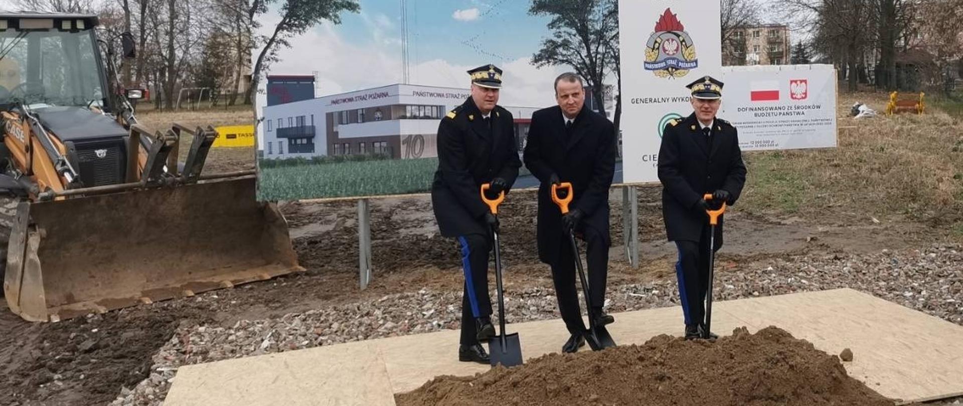 Wielkopolski komendant wojewódzki , wojewoda wielkopolski i komendant główny PSP wbijają szpadle w ziemię, za nimi wizualizacja rozpoczętej inwestycji