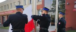 Poczet flagowy przyczepia flagę Polski do masztu w celu uroczystego podniesienia państwowej flagi. W tle znajduje się budynek straży pożarnej oraz niebo