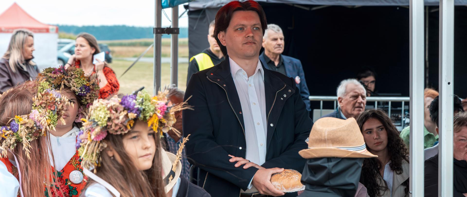 Inspektor WIORiN podczas dożynek w Piotrkosicach trzyma w dłoniach symboliczny bochenek chleba. W tle widoczni są uczestnicy uroczystości, w tym dziewczynki w wiankach z kolorowych kwiatów i kłosów zbóż