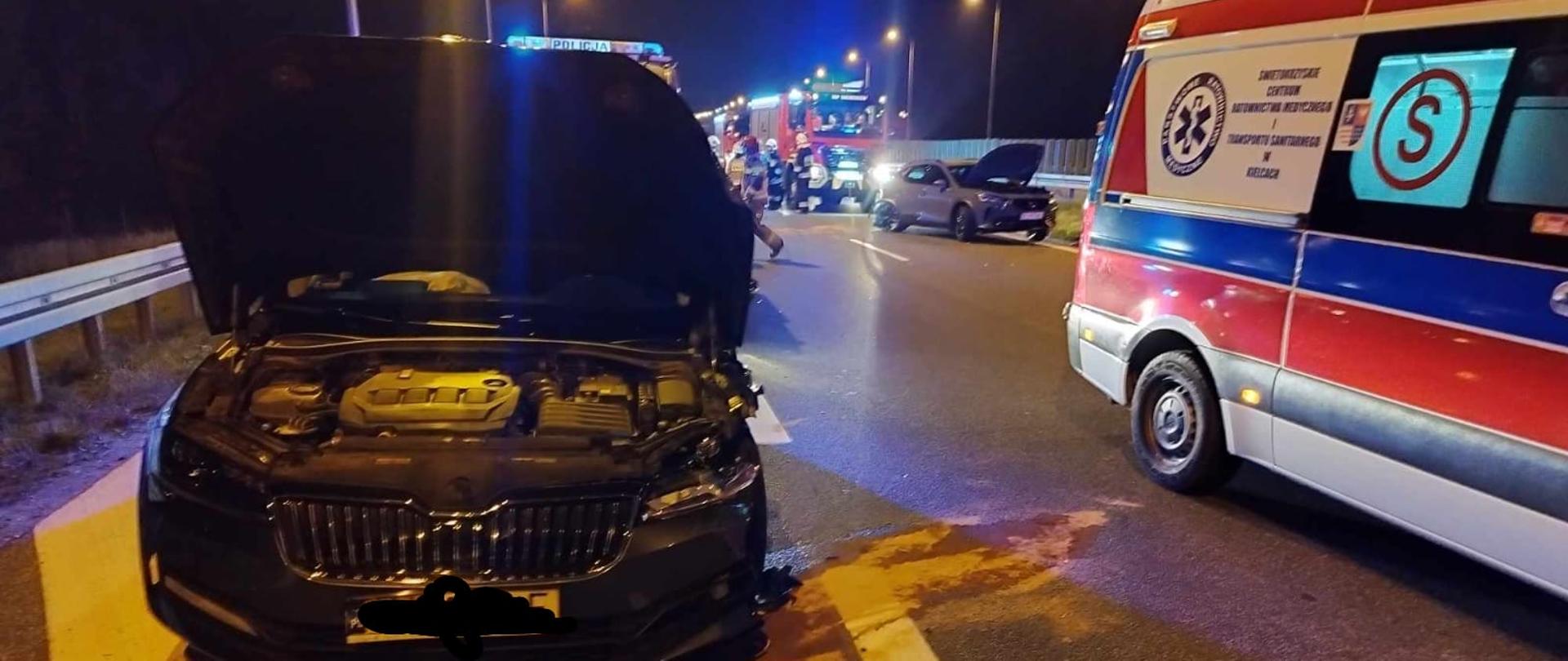 Zdjęcie przedstawia samochód osobowy po wypadku samochodowym, który stoi na poboczu jezdni z podniesioną maską . Obok niego stoi karetka pogotowia i widać wylane płyny eksploatacyjne posypane sorbentem. W tle widać inny samochód uszkodzony i strażaków.