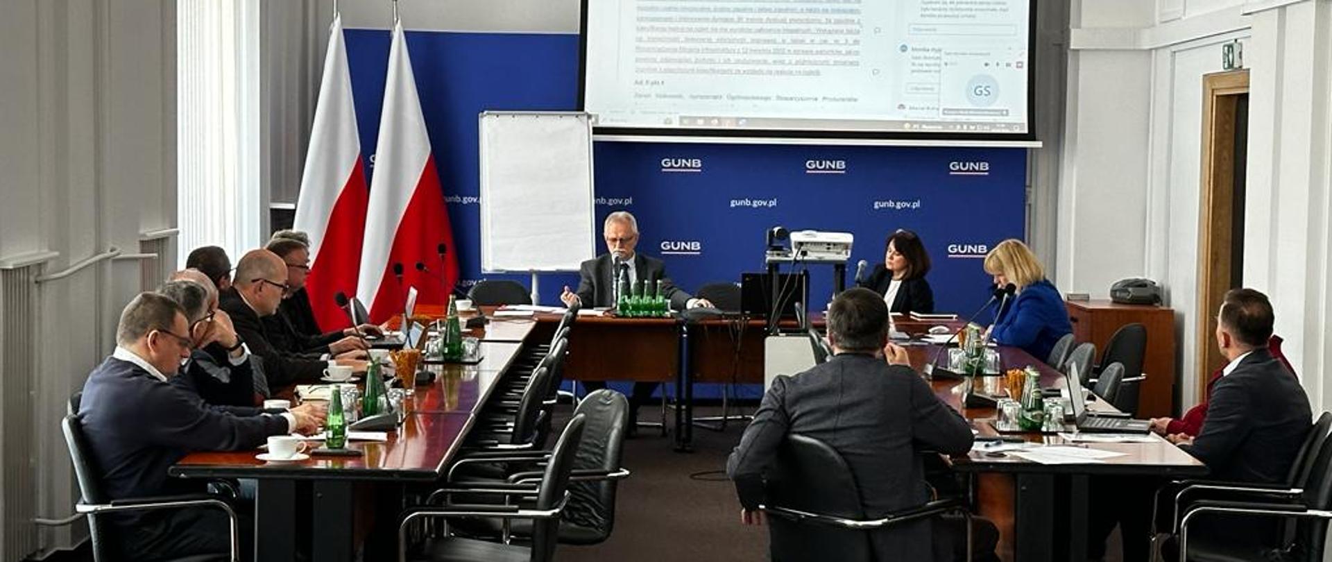 Dwóch pracowników Głównego Urzędu Nadzoru Budowlanego (GUNB) siedzi przy stole konferencyjnym podczas spotkania online; w tle widać trzy flagi Polski, ściankę z logo GUNB oraz tablicę. Na stole znajdują się laptopy, mikrofony, dokumenty i filiżanki.