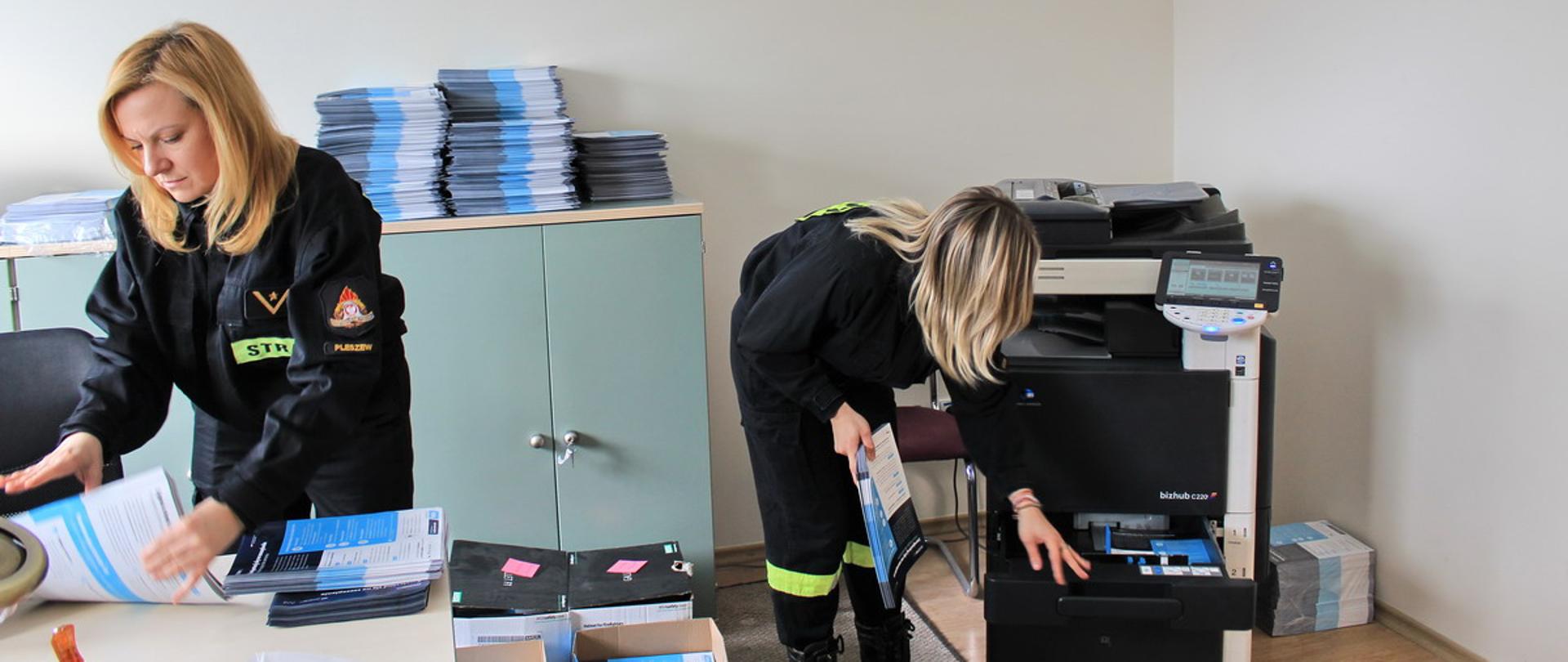 Zdjęcie wykonane w czasie przygotowania ulotek. Dwie funkcjonariuszki PSP przygotowują gotowe ulotki, rozdzielając w kartony dla Jednostek Ochotniczych Straży Pożarnych, które w swoich miejscowościach będą rozdawać je mieszkańcom. 