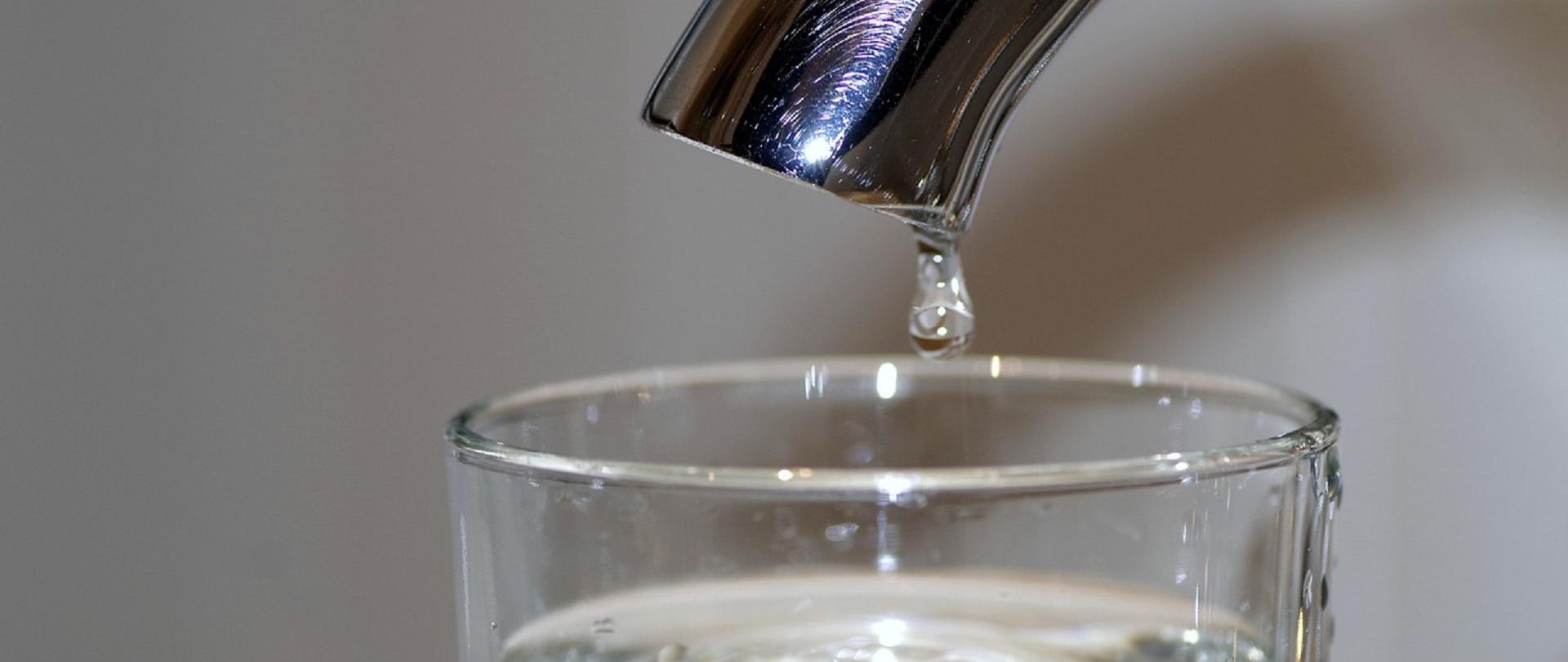 Szklanka wypełniona wodą stojąca pod kranem symbolizująca wodę zdatną do picia 