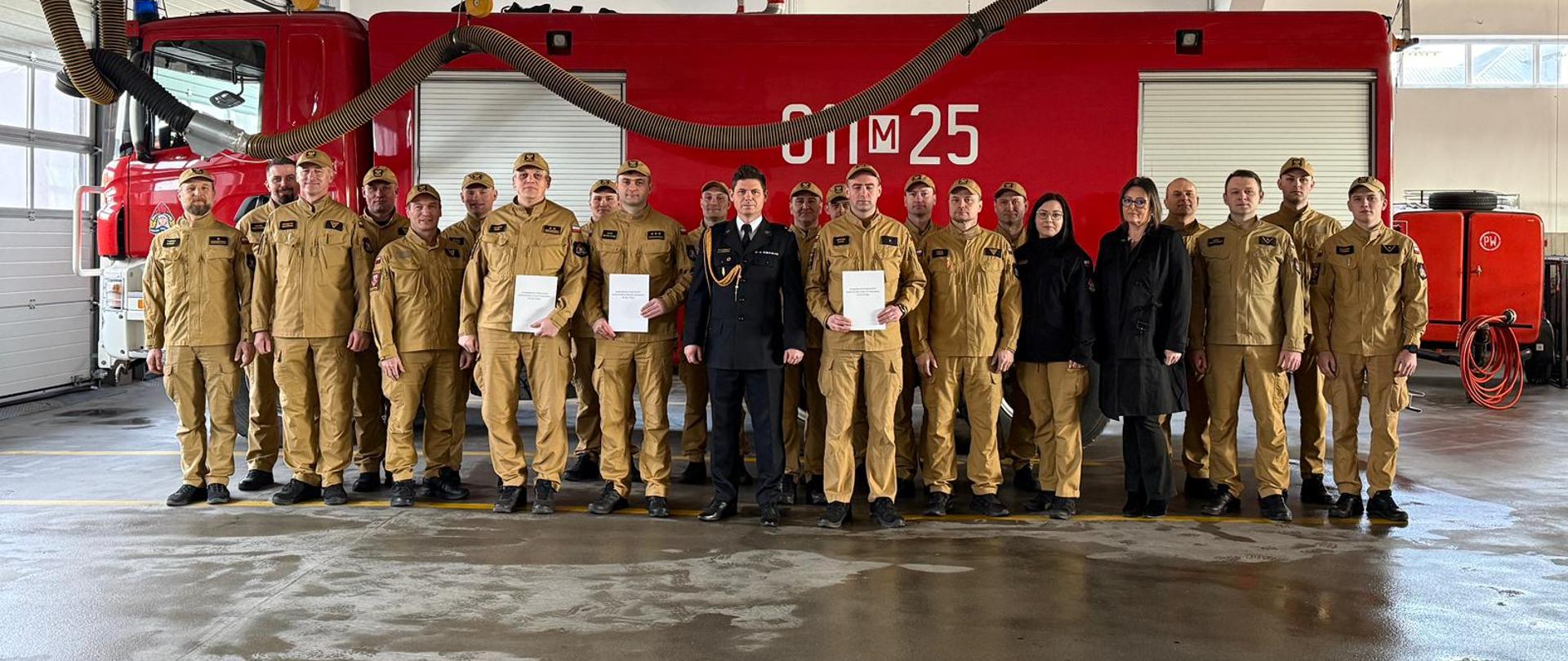 Uroczysta zbiórka strażaków Państwowej Straży Pożarnej w garażu jednostki. Funkcjonariusze w piaskowych mundurach stoją w dwuszeregu przed dużym wozem gaśniczym. Tło stanowi wnętrze przestronnego garażu strażackiego. Bezpośrednio za szeregiem strażaków stoi ciężki, czerwony samochód gaśniczy z widocznym numerem operacyjnym „25” na boku. Do pojazdu podłączone są przewody systemu odciągu spalin zwisające z sufitu. Po lewej stronie znajdują się duże przeszklone bramy garażowe, a na betonowej posadzce widoczne są żółte pasy wyznaczające miejsca postojowe.