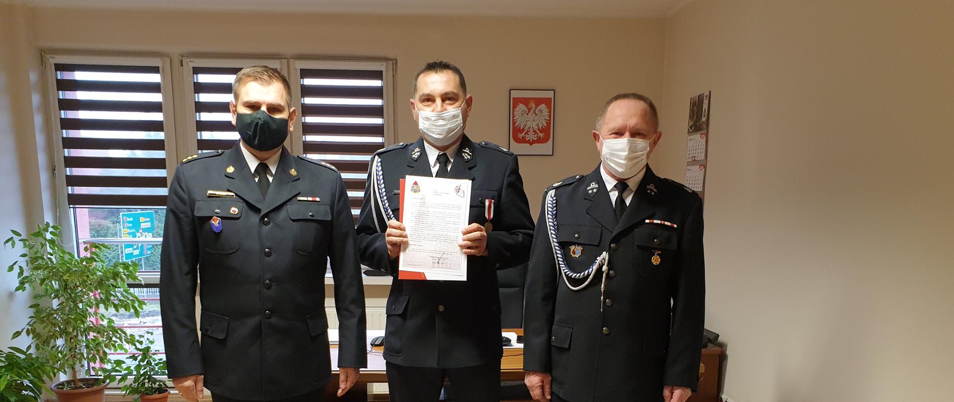 Na zdjęciu widać Z-cę Komendanta PSP w Kaliszu kpt. Damiana Knopa i dwóch druhów z OSP Marcjanów. Za osobami widać okno pomieszczenia, na ścianie godło państwowe. Osoby stoją na szarym dywanie. 