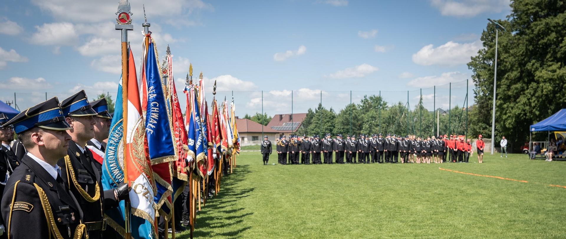 Zdjęcie przedstawia z lewej strony poczty sztandarowe Państwowej Straży Pożarnej i Ochotniczych Straży Pożarnych z terenu powiatu łódzkiego wschodniego. W oddali widoczne pododdziały PSP i OSP w mundurach galowych. Z prawej strony widoczny pododdział Młodzieżowych Drużyn Pożarniczych. 