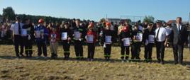 Zawody sportowo-pożarnicze w Jeziorach. Plac ćwiczeń. Na tle drużyn biorących udział w zawodach, ustawionych na zbiórce końcowej, w szeregu stoją dowódcy drużyn w kategorii IM, ze statuetkami, dyplomami i medalami w rękach. Towarzyszy im burmistrz, prezes ZOMG ZOSP RP i komendant gminny.