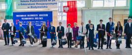 Na sali gimnastycznej stoi rząd elegancko ubranych osób, za nimi ściana z drewnianymi drabinkami przesłonięta granatowym materiałem z białym napisem Podsumowanie wojewódzkich konkursów przedmiotowych 2022/2023, nad nim napis Szkoła Podstawowa nr 24 z oddziałami dwujęzycznymi w Białymstoku, obok polska flaga.