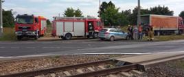 Na zdjęciu widać dwa samochody pożarnicze, radiowóz oraz uszkodzony samochód ciężarowy. Przy radiowozie stoją strażacy, policjanci i pracownicy PKP