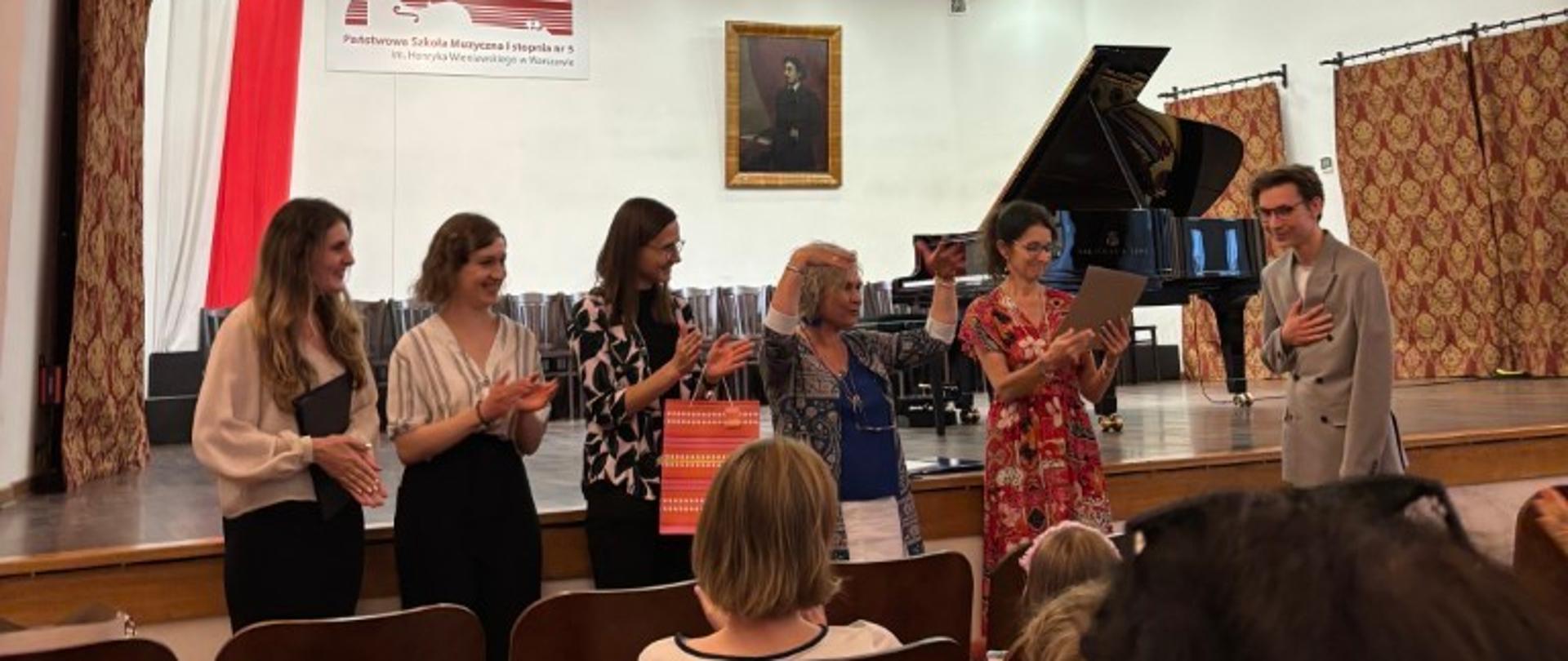 Zdjęcie kolorowe uroczystość na scenie. Sześć osób (pięć kobiet i jeden mężczyzna) stoi obok siebie. Kobieta po prawej w kwiecistej sukience trzyma w rękach dyplomy i nagrody. Obok niej mężczyzna w jasnej marynarce. Reszta kobiet w różnych ubraniach. Wszyscy biją brawo. W tle fortepian, obraz, tablica z napisem „Państwowa Szkoła Muzyczna I stopnia nr 5 im. Witolda Lutosławskiego w Warszawie” i polska flaga. Na dole widoczna publiczność siedząca na krzesłach i bijąca brawo.