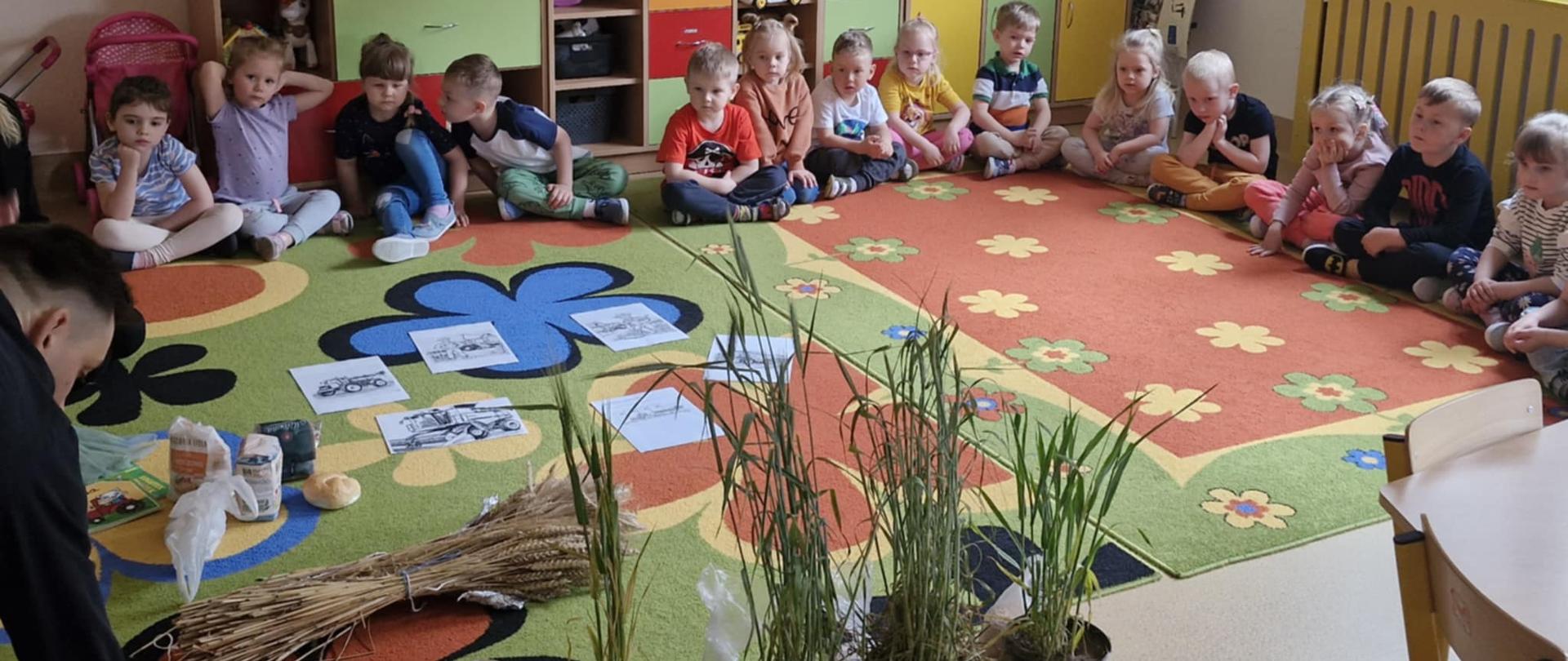 rośliny rolnicze w doniczkach, głównie zboża na pierwszym planie, wokół siedzą dzieci i patrząc w stronę prowadzącego
