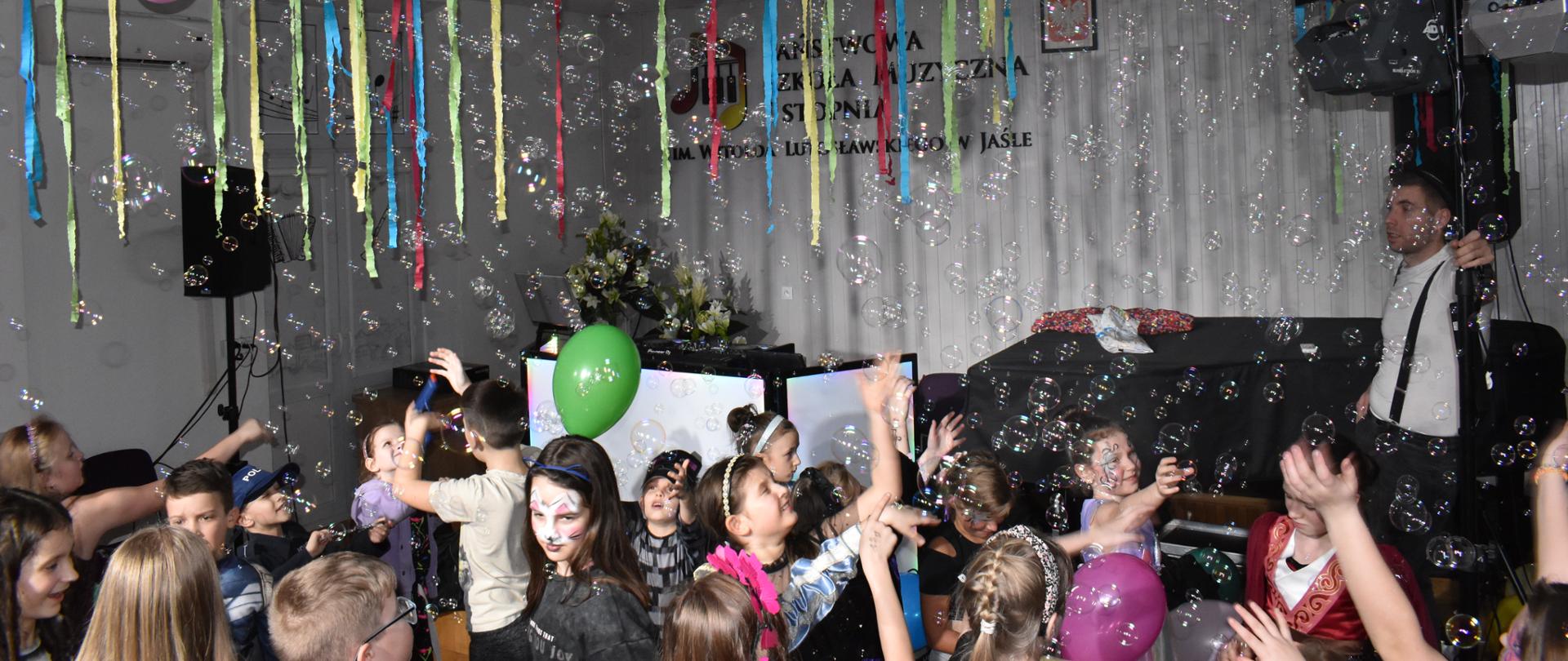 Grupa uczniów tańczących w auli PSM wśród dużej ilości baniek mydlanych. Ogólna kolorystyka – biała i kolorowa.