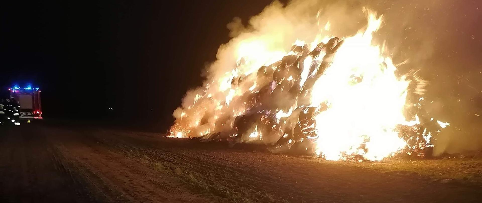 Na polu widać pożar stogu balotów słomy, jest noc, w lewym rogu widać samochód straży pożarnej