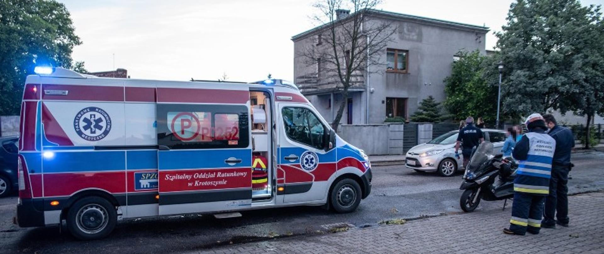 Karetka pogotowia zaparkowana na drodze. W karetce uchylone są drzwi przedziału medycznego oraz włączone sygnały świetlne. Obok pojazdu stoi strażak kierujący akcją, ktory wraz z funkcjonariuszem Policji sporządza dokumentację. Na poboczu drogi zaparkowany jest skuter, który uczestniczył w zdarzeniu. W oddali widać samochód osobowy, który również stoi na drodze z włączonymi światłami postojowymi. Srebrny ford fiesta to również uczestnik zdarzenia drogowego. Obok pojazdu stoi drugi funkcjonariusz Policji, który prowadzi czynności dochodzeniowe. 